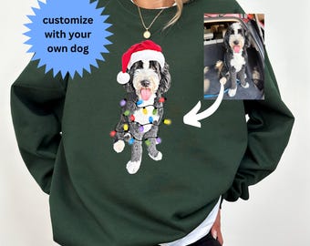 May include: A dark green sweatshirt with a white dog wearing a Santa hat and Christmas lights printed on the front. The dog is surrounded by a white border. The sweatshirt has a crew neck and long sleeves. The text "customize with your own dog" is in a blue starburst shape in the top left corner.