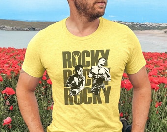 May include: A man wearing a yellow t-shirt with a graphic of two boxers and the text "Rocky Balboa Rocky". He is standing in a field of red poppies.