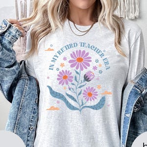 May include: A light gray t-shirt with the text "In My Retired Teacher Era" in a circle around a floral design with purple flowers and green leaves.
