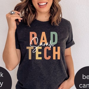 Könnte beinhalten: Ein dunkelgraues T-Shirt mit dem Text "RAD Sarah TECH" in bunten Blockbuchstaben. Die Buchstaben sind in den Farben Grün, Gelb, Orange und Rot.
