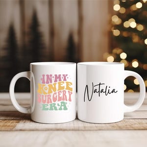 Könnte beinhalten: Zwei weiße Keramikbecher. Auf einem Becher steht in Retrofarben der Text "IN MY KNEE SURGERY ERA". Der andere Becher trägt den Namen "Natalia" in schwarzer Schrift. Beide Becher haben einen weißen Henkel.