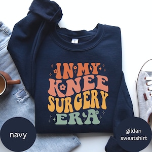 May include: Navy blue Gildan sweatshirt with the text "In My Knee Surgery Era" in a retro, colorful font.
