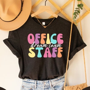 Könnte beinhalten: Ein schwarzes T-Shirt mit dem Text "OFFICE dream team STAFF" in bunten, Retro-Lettern.
