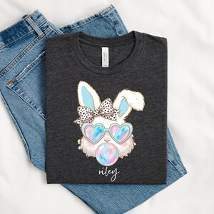 May include: Dark grey t-shirt with a cartoon bunny wearing heart-shaped sunglasses and blowing a bubble. The bunny has a bow and the name "riley" is written below. The shirt is folded on top of blue denim jeans.