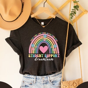 Può includere: Una maglietta nera con un design arcobaleno e cuore. Il testo sulla maglietta recita "Teach them, love them, watch them grow. Student Support Dream Team."