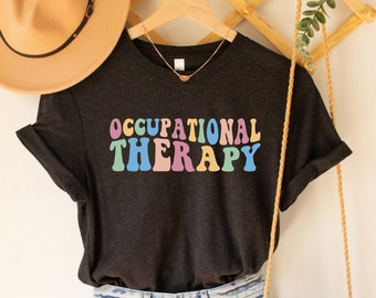 Op de afbeelding: Een zwart T-shirt met de tekst "OCCUPATIONAL THERAPY" in regenboogkleuren. Het shirt hangt aan een houten rek met een bruine hoed en een klein bruin tasje.