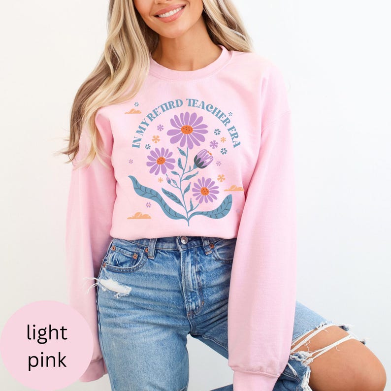 May include: A light pink sweatshirt with the text "In My Retired Teacher Era" and a floral design. The flowers are purple, pink, and blue.