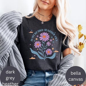 May include: A dark heather grey t-shirt with the text "In My Retired Teacher Era" and a floral design in shades of purple, pink, and blue.