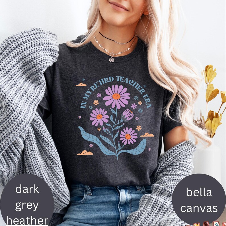 May include: A dark heather grey t-shirt with the text "In My Retired Teacher Era" and a floral design in shades of purple, pink, and blue.