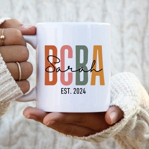 Peut inclure: Une tasse en céramique blanche avec le texte "BCBA Sarah EST. 2024" en lettres majuscules colorées. Les lettres sont orange, rose, verte et jaune.