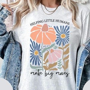 May include: A pale grey sweatshirt featuring a floral design in orange, blue, and pink. The text "HELPING LITTLE HUMANS" is above the flowers, and "make big moves" is below. The sweatshirt has long sleeves.