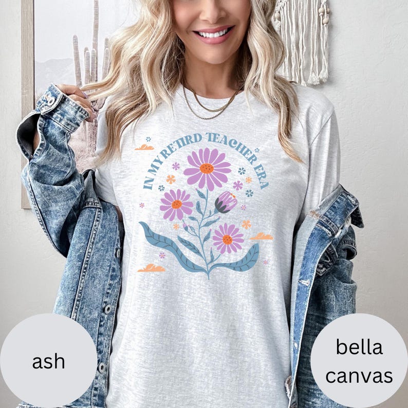 May include: A light gray t-shirt with the text "In My Retired Teacher Era" in a circle around a floral design with purple flowers and green leaves.