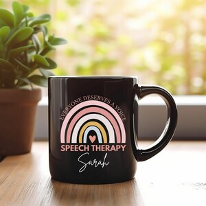Peut inclure: Une tasse à café en céramique noire avec un motif arc-en-ciel et le texte "Everyone deserves a voice" et "Speech Therapy". Le nom "Sarah" est écrit en cursive sous le texte.