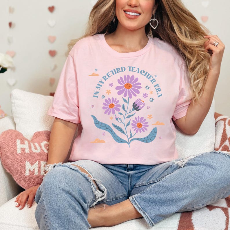 May include: A pink t-shirt with a floral design and the text "In My Retired Teacher Era".