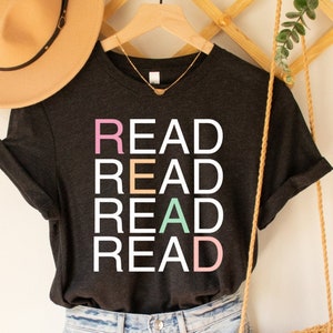 May include: A dark grey t-shirt with the word "READ" repeated four times in white letters, with each letter in a different pastel colour. The shirt is hanging on a wooden hanger.