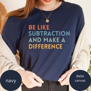 May include: Navy blue t-shirt with the text "BE LIKE SUBTRACTION AND MAKE A DIFFERENCE" in orange, teal, and yellow. The shirt is a Bella Canvas brand.