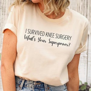 Puede incluir: Una camiseta de color crema con el texto "I survived knee surgery What's your superpower?" impreso en negro.