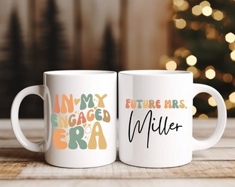 Könnte beinhalten: Zwei weiße Keramikbecher mit einem bunten Design. Der Becher links sagt "In My Engaged Era" mit einem Herzen und einem Ring. Der Becher rechts sagt "Future Mrs. Miller".