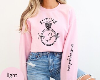 May include: Light pink sweatshirt with the text "FUTURE Mrs. Smith" and a diamond ring graphic. The sleeve has the text "IN MY engaged ERA".