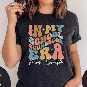 May include: A dark grey t-shirt with a colorful retro design that says "In My School Counselor Era" with the name "Mrs. Smith" underneath.