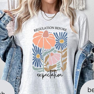 Puede incluir: Camiseta gris claro con un diseño floral. Las palabras "REGULATION BEFORE" están arriba y "expectation" debajo de las flores. Las flores son melocotón, azules y rosas, con acentos verdes y marrones. Prenda de vestir informal.