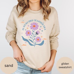 May include: A sand-colored Gildan sweatshirt with the text "In My Retired Teacher Era" in a blue circle with purple flowers.