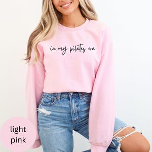 Pu&ograve; includere: Felpa rosa chiaro con la scritta "in my pilates era" in corsivo nero. La felpa girocollo &egrave; abbinata a jeans blu. Le parole "rosa chiaro" sono in un cerchio rosa in basso a sinistra.