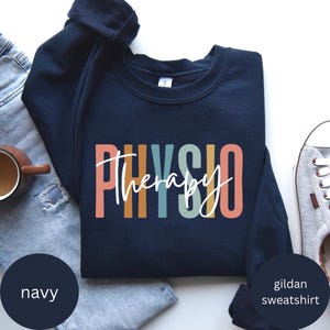 May include: Navy blue sweatshirt featuring the word "PHYSIO" in colourful block letters and the word "therapy" in a cursive font. The sweatshirt is a Gildan brand.