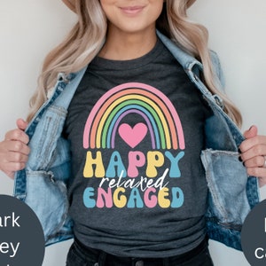 以下が含まれることがあります： 虹とピンクのハートが描かれたダークグレーのTシャツ。"HAPPY relaxed ENGAGED"というテキストがレトロなフォントでプリントされています。