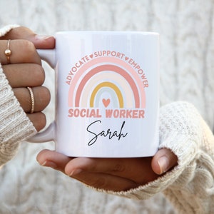 Könnte beinhalten: Eine weiße Keramiktasse mit einem rosafarbenen, gelben und weißen Regenbogenmuster. Der Regenbogen ist von den Worten "Advocate", "Support", "Empower" umgeben. Der Text "Social Worker" befindet sich unter dem Regenbogen, und der Name "Sarah" ist darunter in Kursivschrift geschrieben.