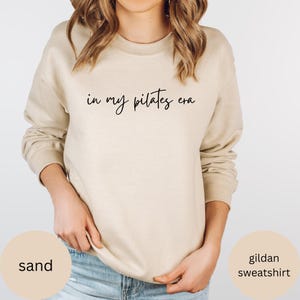 Pu&ograve; includere: Felpa Gildan color sabbia con la scritta "in my pilates era" in corsivo nero. La felpa ha girocollo e maniche lunghe. L'immagine include anche le parole "sand" e "gildan sweatshirt".