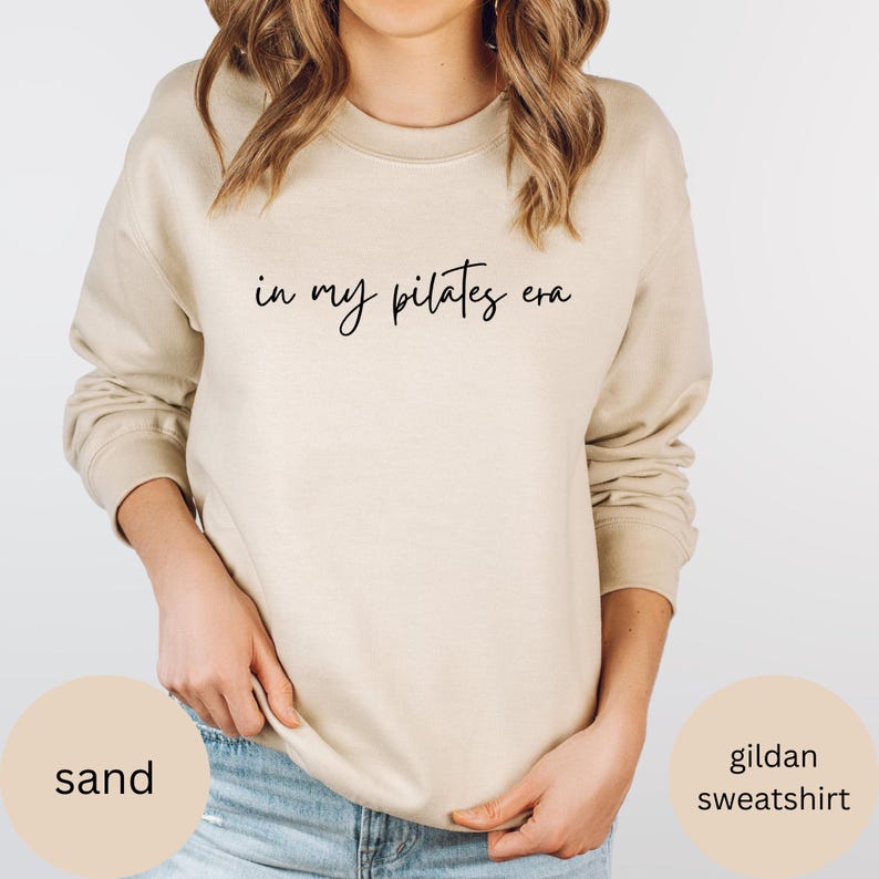 Pu&ograve; includere: Felpa Gildan color sabbia con la scritta "in my pilates era" in corsivo nero. La felpa ha girocollo e maniche lunghe. L'immagine include anche le parole "sand" e "gildan sweatshirt".