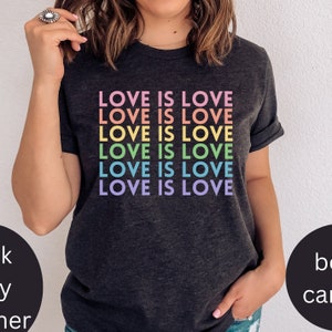 Puede incluir: Una camiseta gris oscuro con la frase "LOVE IS LOVE" repetida seis veces en colores arcoíris.