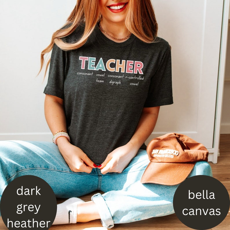 May include: A dark heather grey t-shirt with the word "TEACHER" written in colorful letters. The word is defined with the words "consonant", "vowel", "consonant", "r-controlled", "team", "digraph", and "vowel".