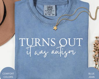 May include: A blue jean coloured t-shirt with the text "TURNS OUT it was autism" in white. A gold necklace with a star pendant is on the shirt. The shirt has a Comfort Colors tag.