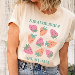 Puede incluir: Una camiseta color crema con un diseño de fresas. El diseño incluye fresas rosas y naranjas con tallos verdes, y el texto "STRAWBERRIES ARE MY JAM" en letras turquesas. La camiseta tiene mangas enrolladas.