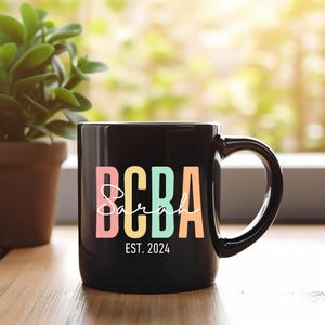 Op de afbeelding: Zwarte koffiemok met een kleurrijk ontwerp "BCBA Sarah EST. 2024".
