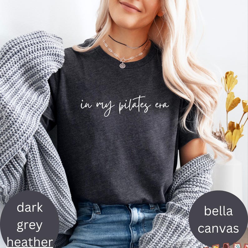 Pu&ograve; includere: T-shirt grigio scuro m&eacute;lange con la scritta "in my pilates era" in corsivo bianco. La maglietta &egrave; in materiale Bella Canvas. La modella indossa un maglione grigio.