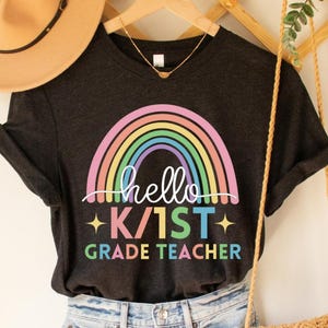May include: Dark grey t-shirt featuring a rainbow design and the text "hello K/1ST GRADE TEACHER" in various colours. The t-shirt is displayed on a wooden hanger.