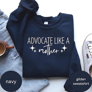 Peut inclure: Sweat-shirt bleu marine avec l'inscription blanche "ADVOCATE LIKE A mother". Le sweat-shirt est de la marque Gildan. L'image montre également un jean bleu, une tasse à café et une basket.