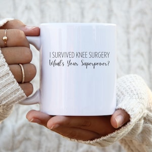 Könnte beinhalten: Weißer Keramikbecher mit dem Text "I Survived Knee Surgery What's Your Superpower?" in Schwarz. Der Becher wird von einer Person gehalten, die einen weißen Pullover trägt.