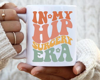 May include: White ceramic mug with the text "IN MY HIP SURGERY ERA" in orange, peach, yellow, and green retro font. The mug has a white handle and is held by a person wearing a white sweater.