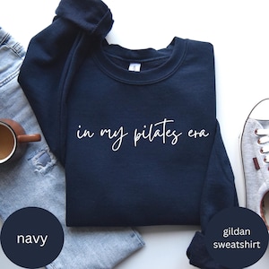 Pu&ograve; includere: Felpa blu navy con la scritta bianca "in my pilates era". La felpa &egrave; del marchio Gildan. L'immagine mostra anche un paio di jeans blu, una tazza da caff&egrave; e una sneaker grigia.