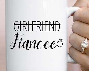 Op de afbeelding: Witte keramische mok met zwarte tekst die "Girlfriend" met een streep erdoor en "Fiancee" met een diamanten ring symbool leest.