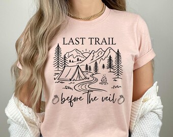 May include: Pink t-shirt featuring a black graphic of a tent in a mountain landscape. The text "Last Trail Before The Veil" is printed below the graphic.