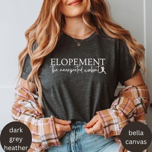 Può includere: T-shirt grigio scuro melange con la scritta bianca "ELOPEMENT The unexpected workout". La maglietta è realizzata in materiale Bella Canvas e presenta una grafica di una persona che corre. La modella indossa una camicia a quadri.