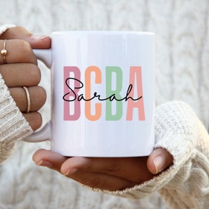 Op de afbeelding: Een witte keramische mok met de tekst "BCBA Sarah" in kleurrijke letters. De letters zijn roze, oranje, groen en roze.