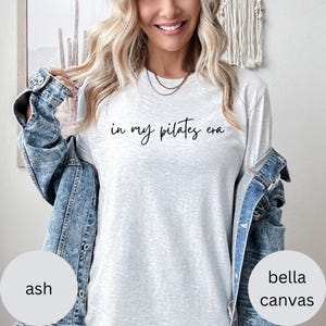 Pu&ograve; includere: Maglietta a maniche lunghe grigio chiaro con la scritta "in my pilates era" in corsivo nero. La maglietta &egrave; color cenere e realizzata in materiale Bella Canvas. La persona indossa una giacca di jeans.