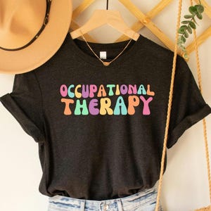 May include: A dark gray t-shirt with the text "Occupational Therapy" in colorful, retro-style lettering.