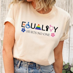 Peut inclure: Un t-shirt crème avec le mot "EQUALITY" écrit avec différents symboles représentant différents groupes de personnes. Les symboles incluent un triangle arc-en-ciel, un poing, un fauteuil roulant, un symbole transgenre et une fleur. Le texte "HURTS NO ONE" est imprimé sous le mot "EQUALITY".